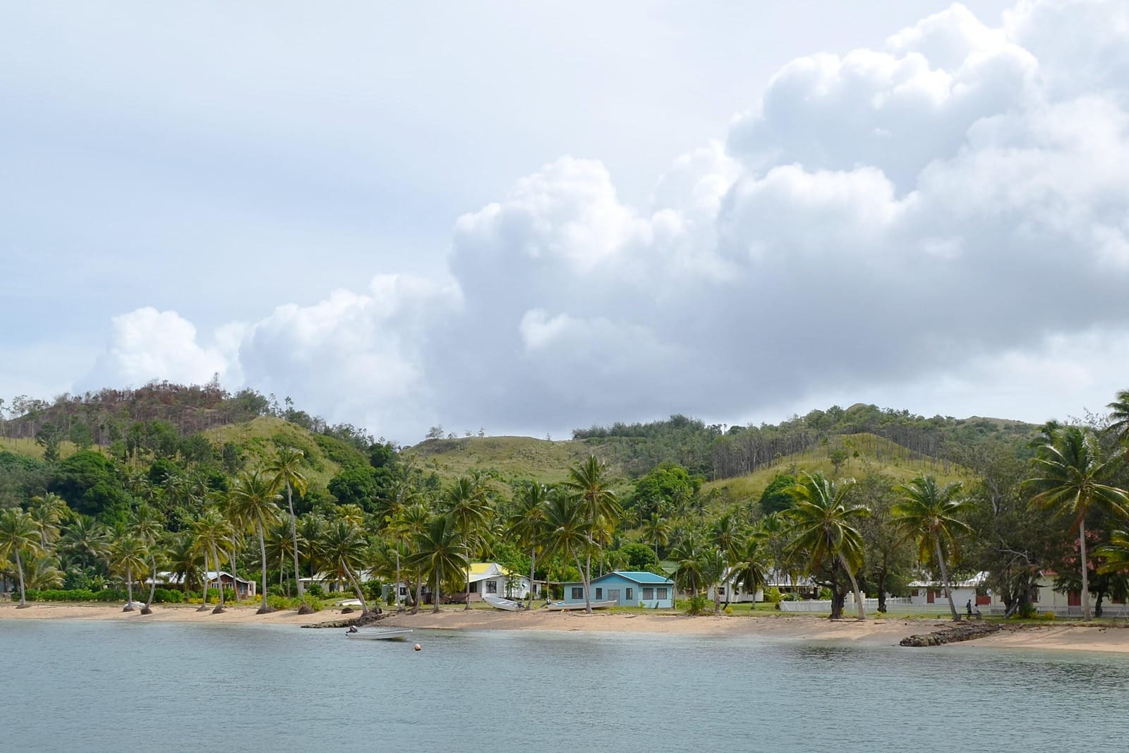 Palm trees, small houses and boats pulled up on a sandy beach beside calm water, green hills behind