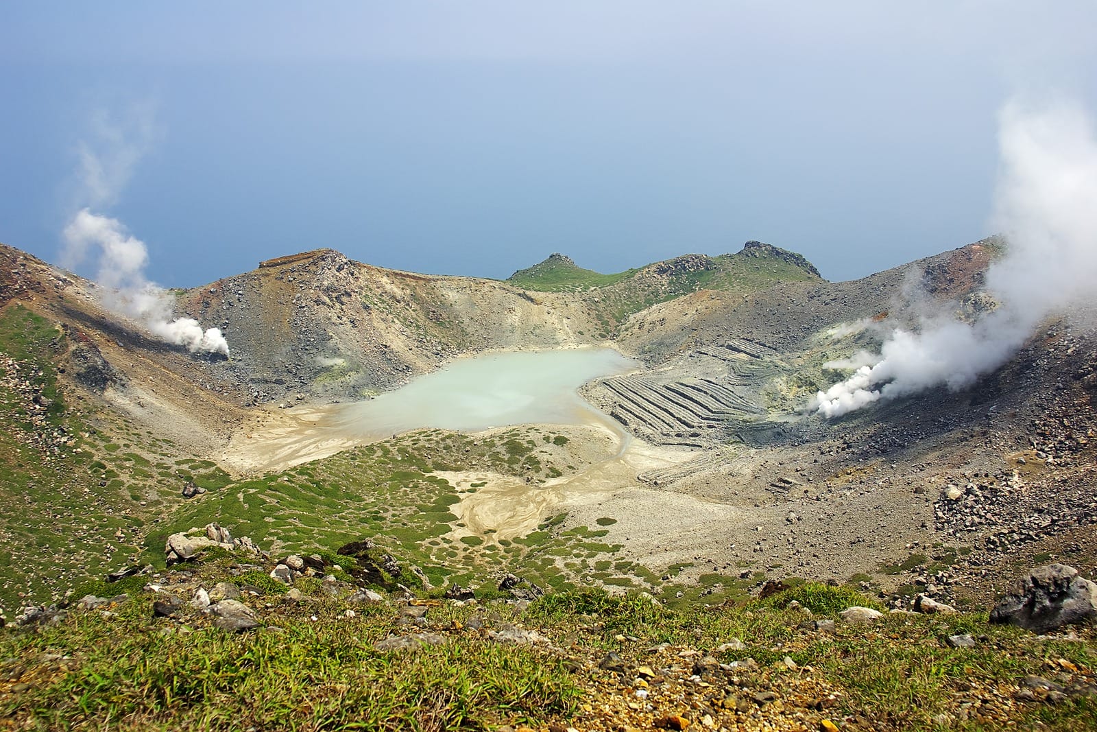 A steaming volcanic crater with a pale lake in it, green slopes in front and the sea beyond