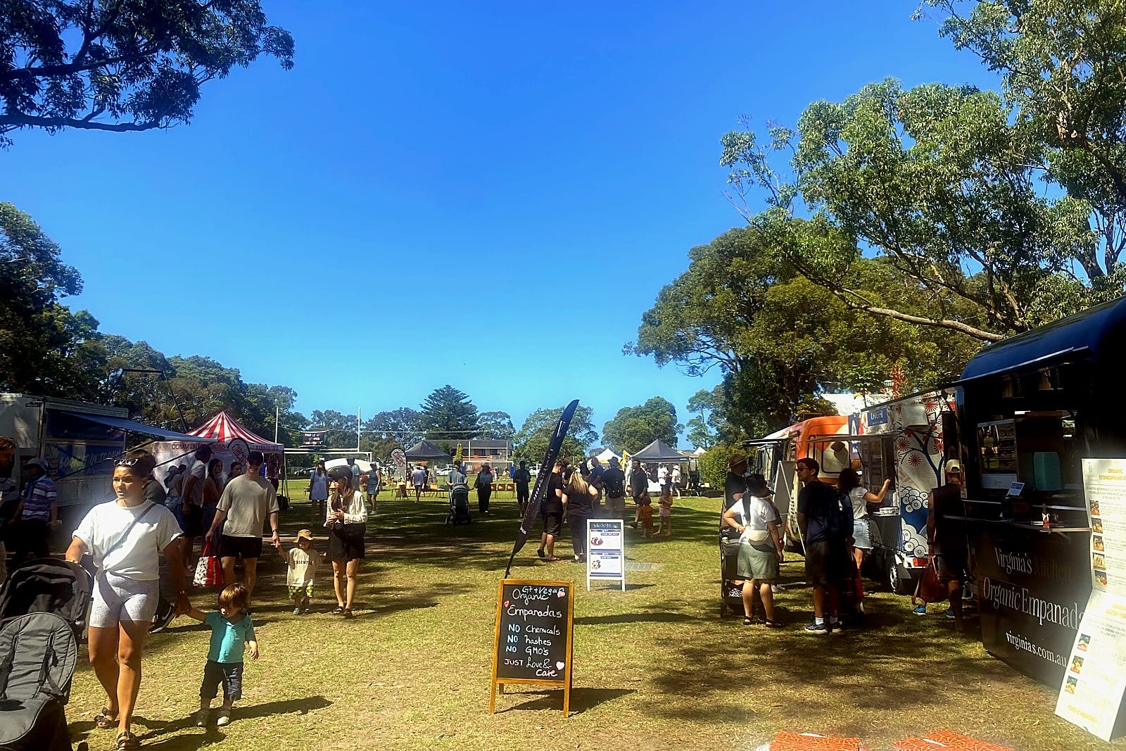Festival goers on a green field between food vans and trees under a blue sky