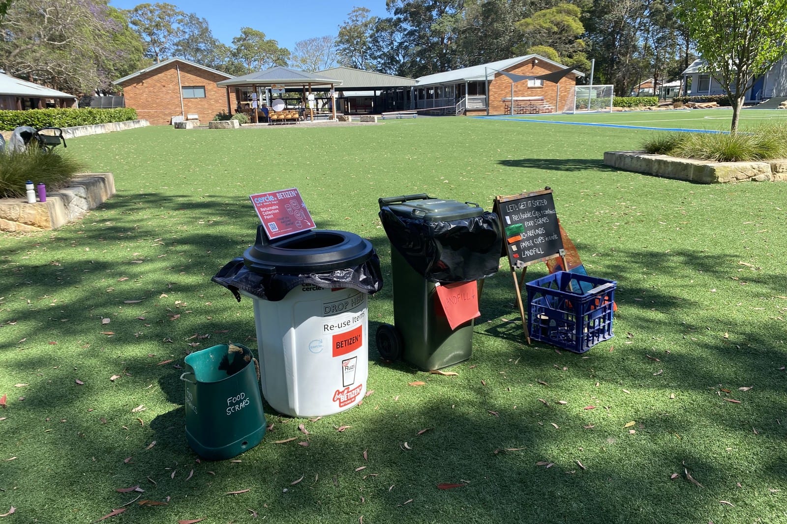 A green food scraps caddy, a white re use bin, a green landfill bin and a chalkboard on a school field