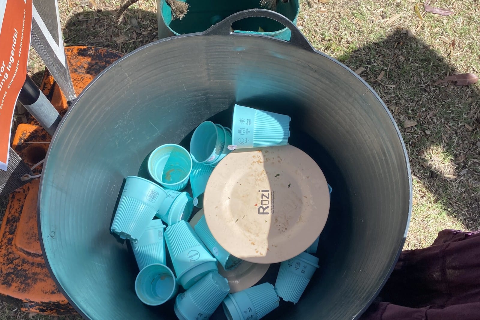 Blue reusable cups and a beige plate in a grey tub on the grass