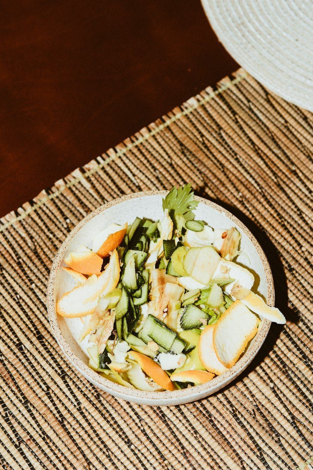 A shallow bowl of vegetable peelings and scraps on a woven table mat