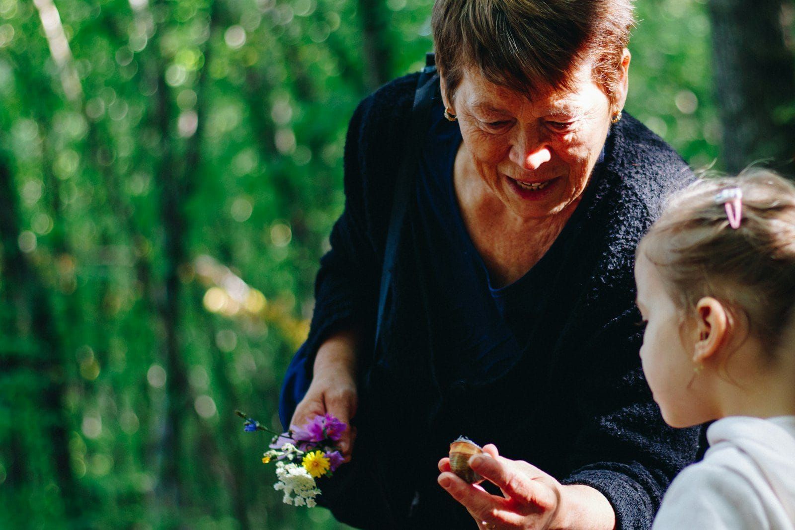 A grandmother in a green wood showing a small girl a posy of wildflowers and a snail