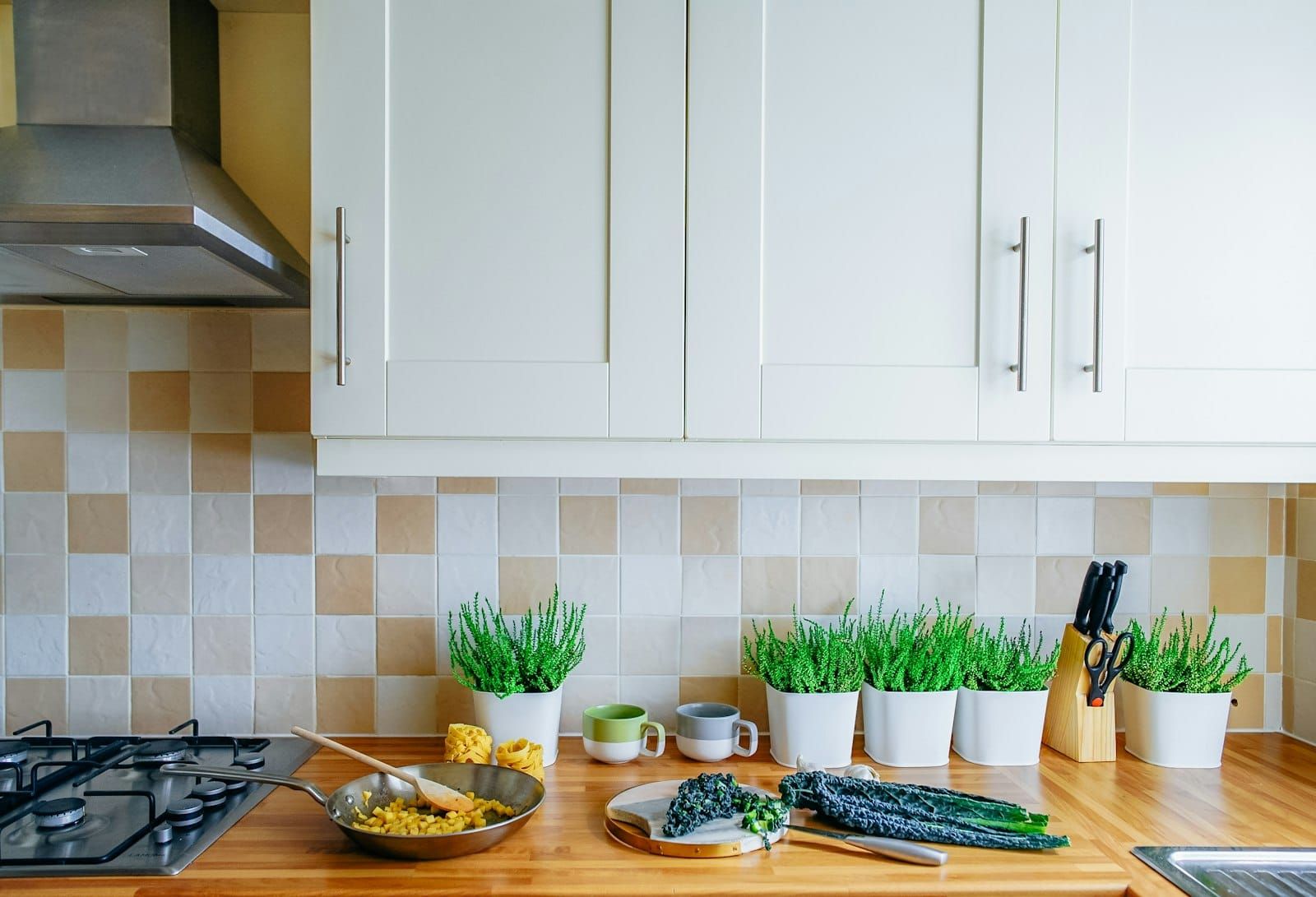 A sunlit kitchen bench with a pan of vegetables cooking, fresh greens and herbs beside it
