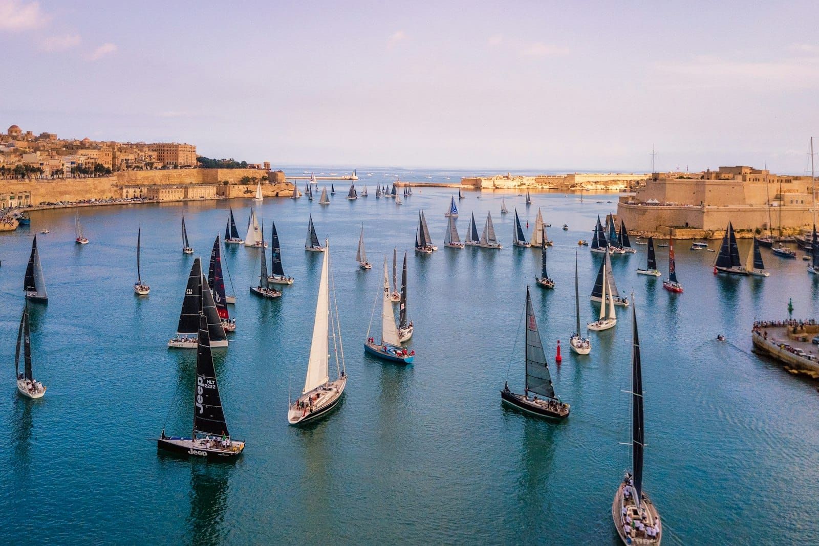 Sailing boats on a calm Mediterranean harbour below honey coloured limestone fortifications