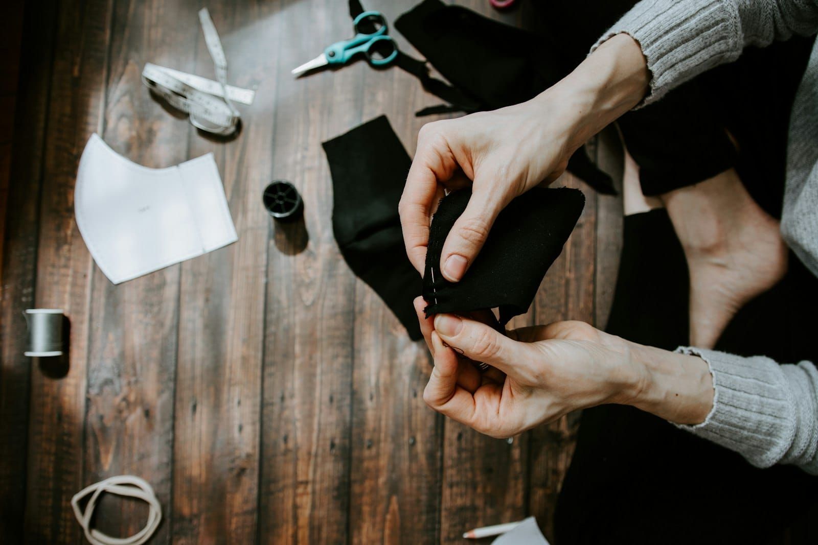 A pair of hands mending dark fabric, with scissors and thread on the floor nearby
