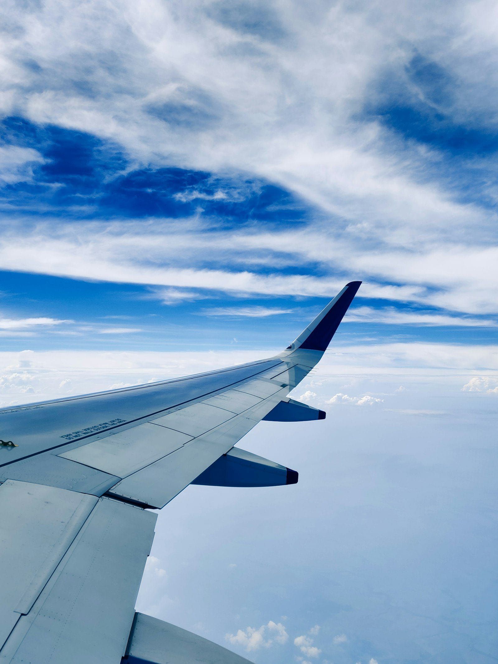 An aircraft wingtip climbing through a bright blue sky with high white clouds