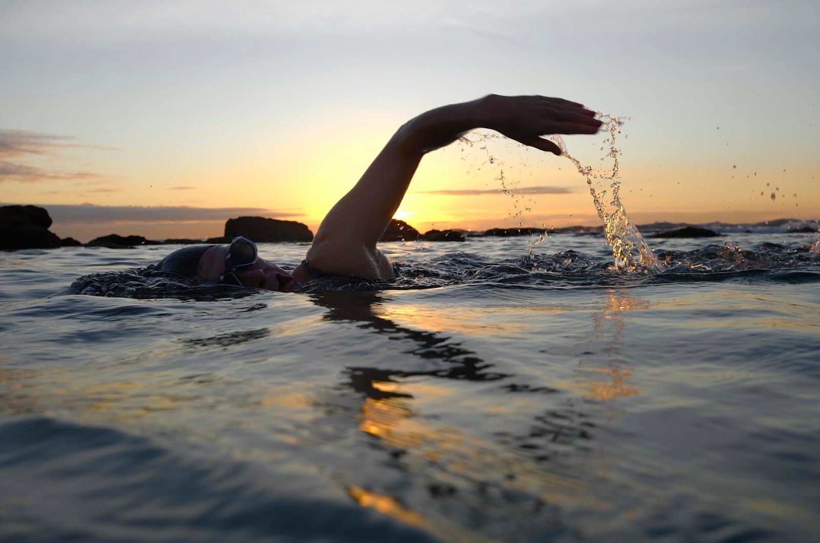 A swimmer in the sea at sunrise, one arm lifted mid stroke against a golden sky.