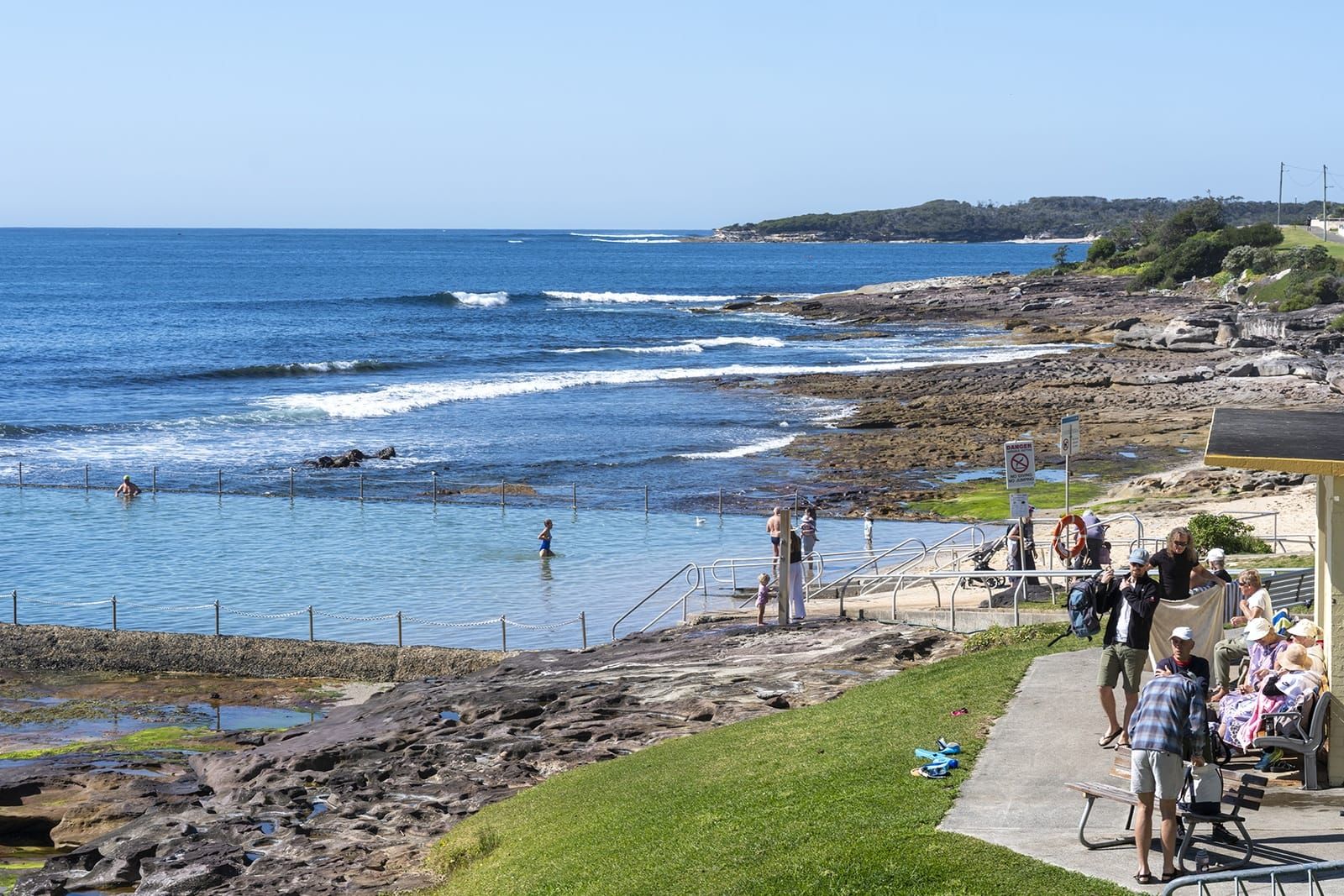 Swimmers in Shelly Park Ocean Pool at Cronulla, with the lawn, the rock shelf and the headland beyond