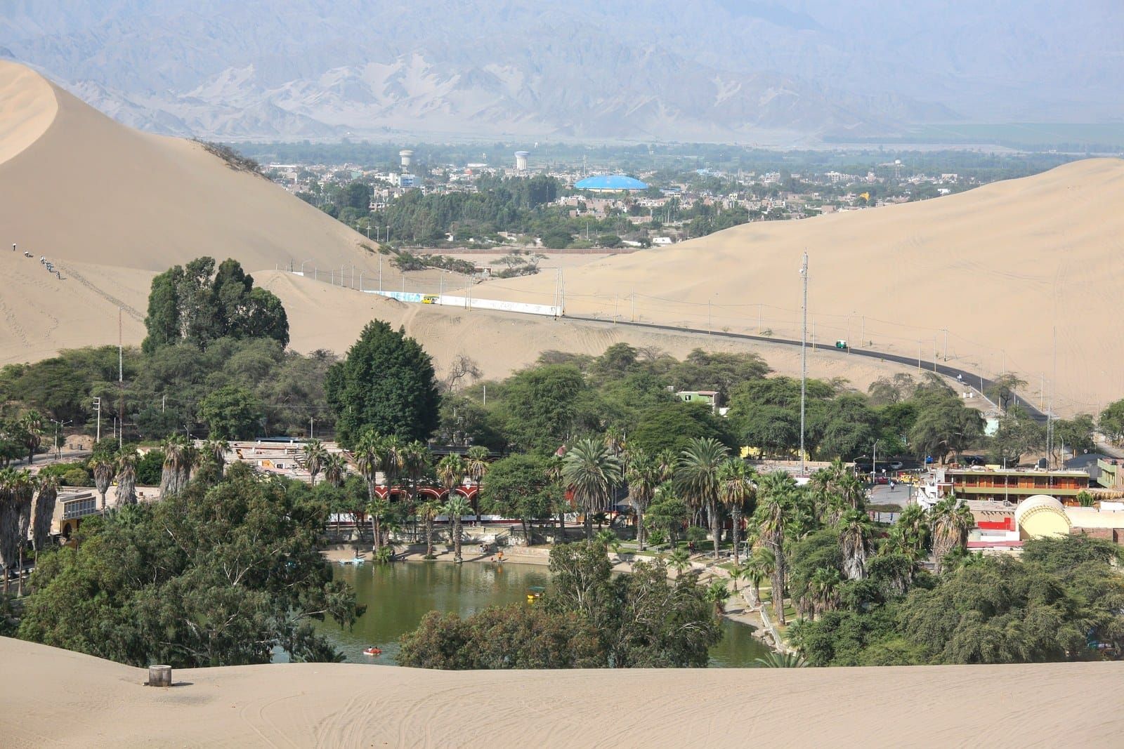 A green lagoon ringed by palms and buildings in a bowl of pale sand dunes, with a city beyond