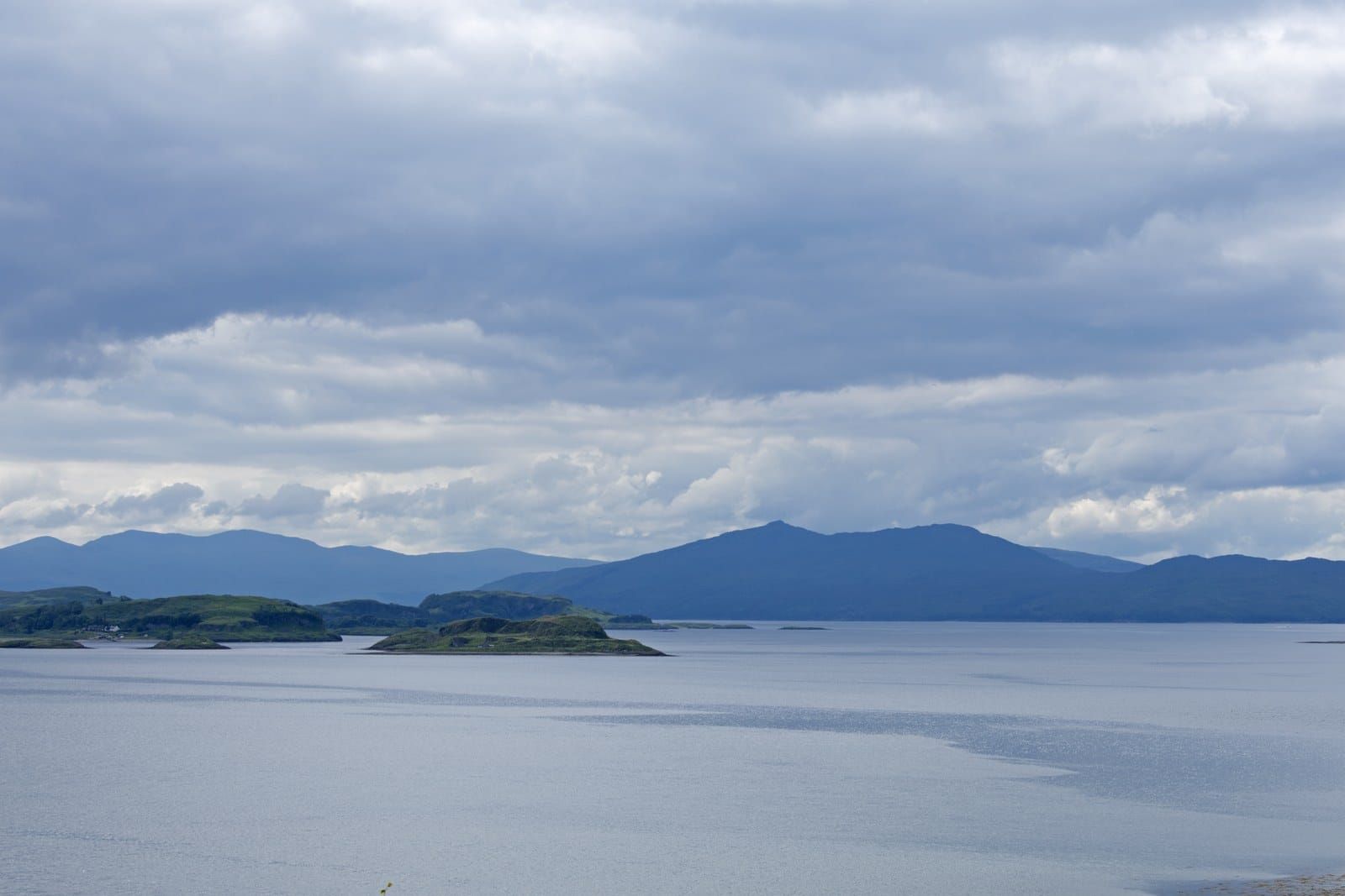 A long low green island under grey cloud across calm sea loch water, hills behind