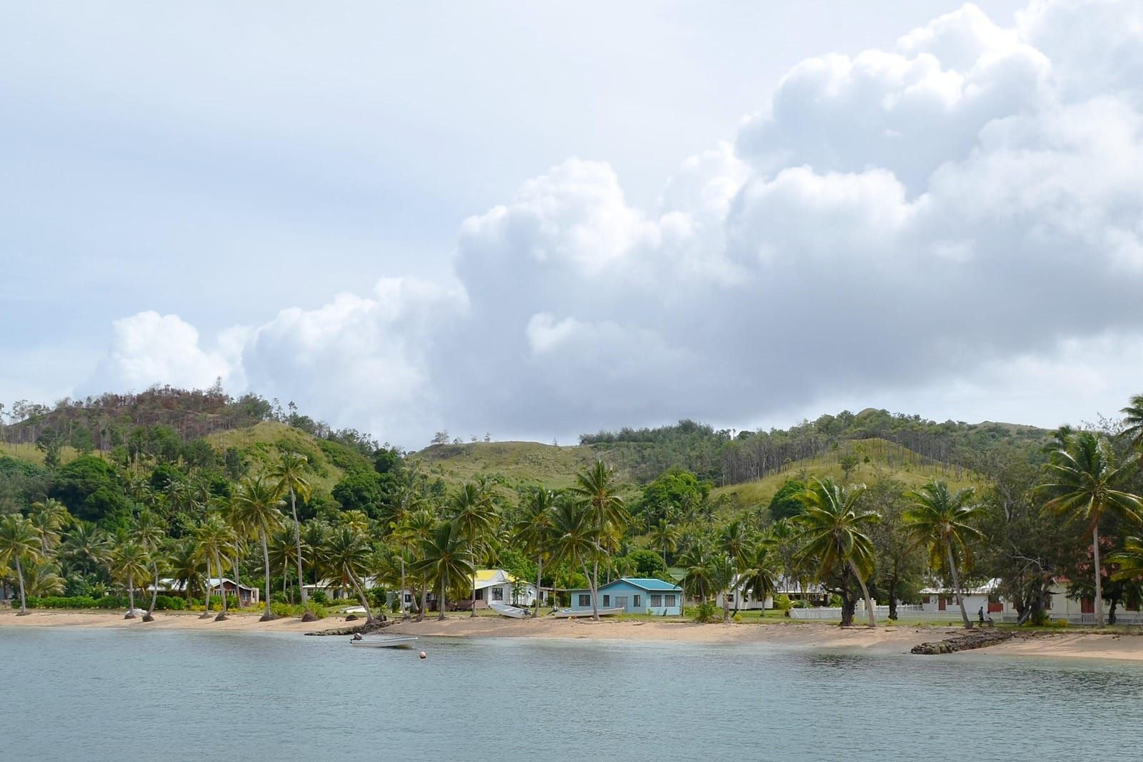 Palm trees, small houses and boats pulled up on a sandy beach beside calm water, green hills behind