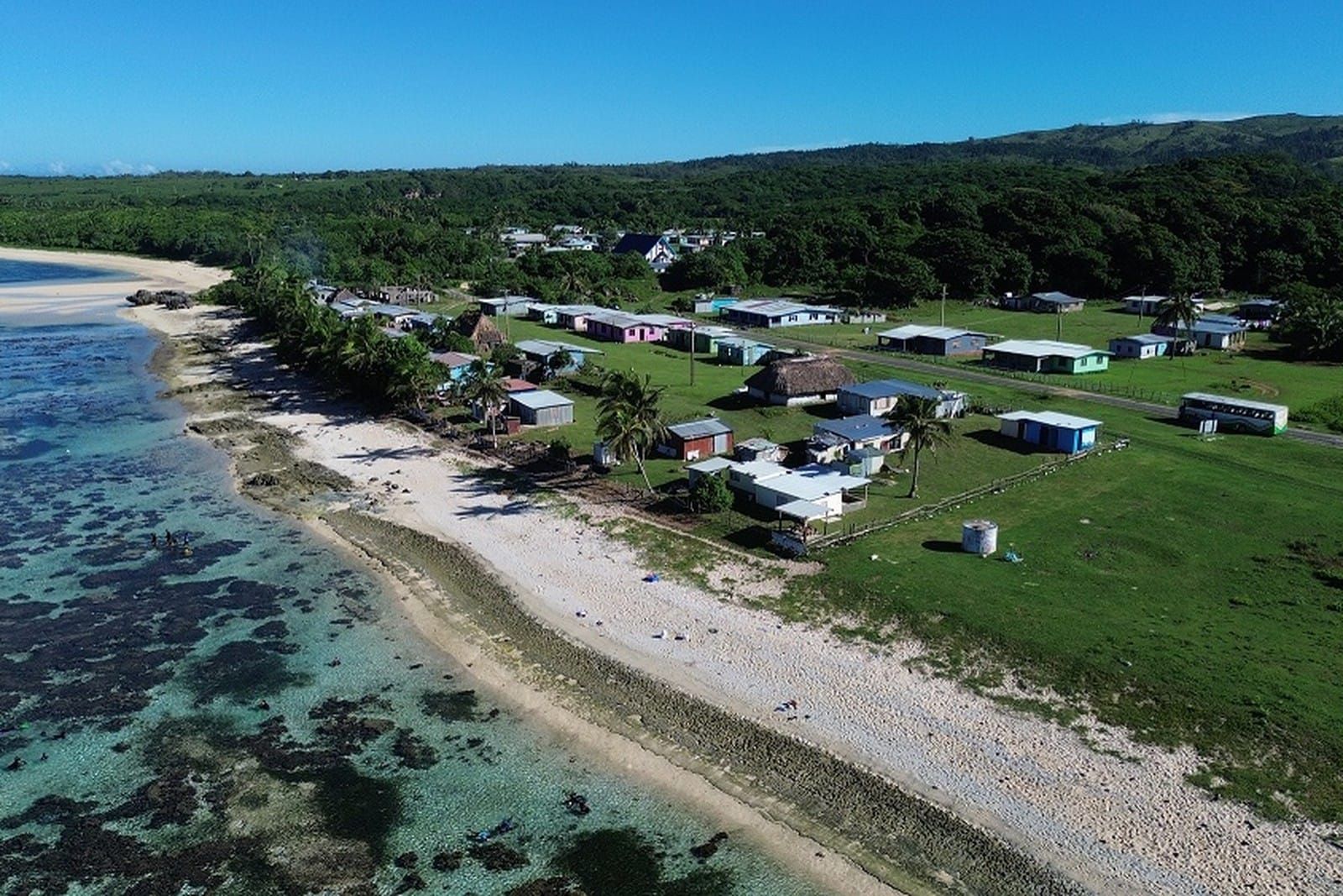 Aerial view of Naidiri village on the Coral Coast of Fiji: tin roofed houses and a thatched bure on green grass beside a white beach, with the reef flat and lagoon in the foreground