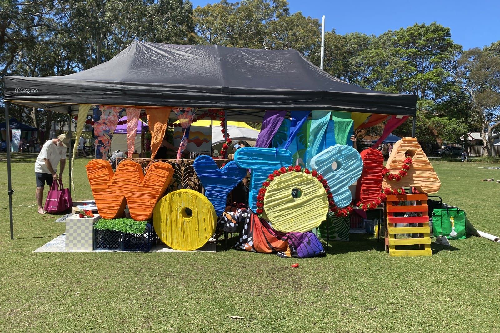 Giant painted letters spelling Woytopia in orange, yellow, blue and pink under a black marquee on a school field