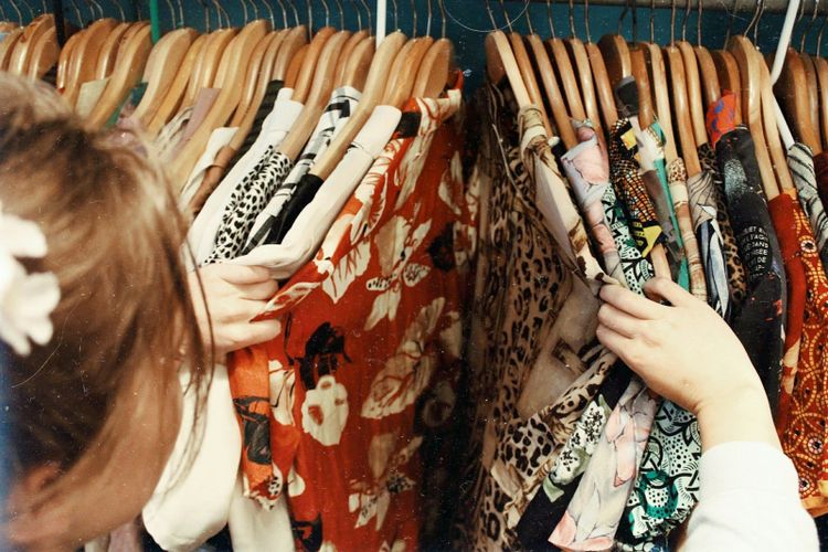 A young woman looking through a crowded rail of colourful secondhand clothes on wooden hangers