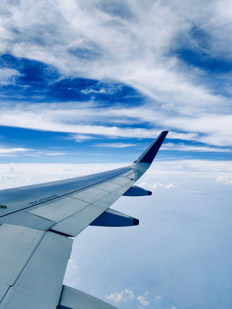 An aircraft wingtip climbing through a bright blue sky with high white clouds