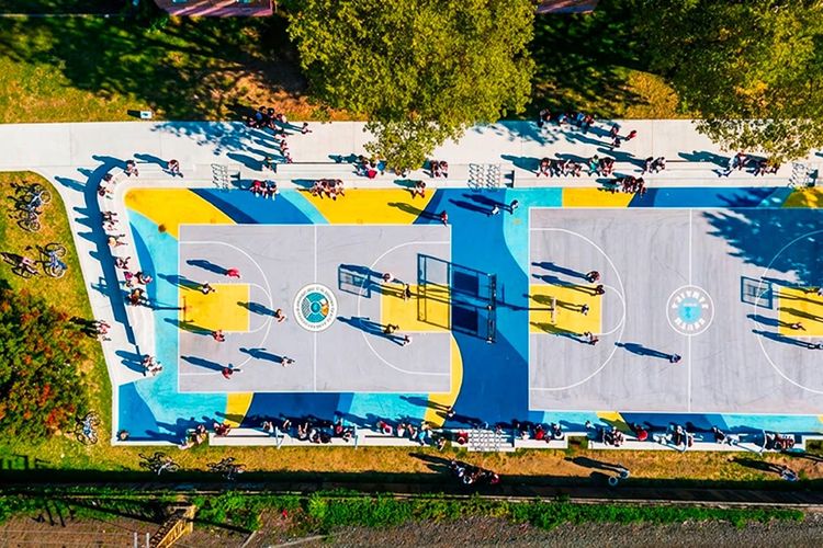 Aerial view of two basketball courts painted blue and gold at the South Jamaica Houses in Queens, people on the paths around them and a rail line alongside