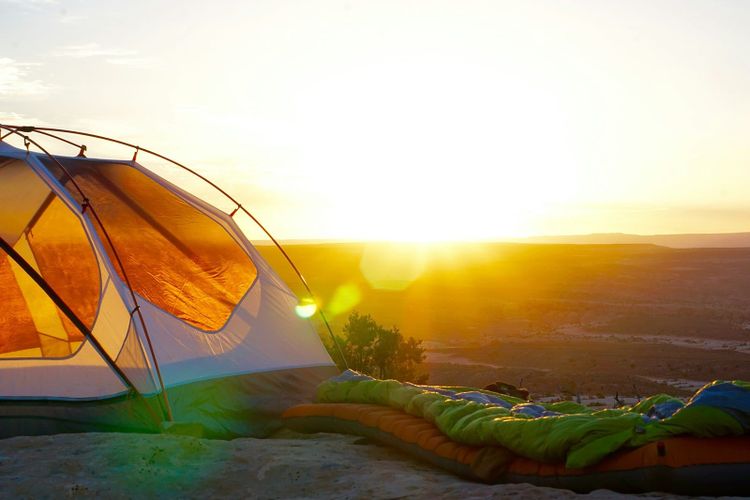 A tent glowing in early sunlight with sleeping bags laid out beside it, looking over a wide valley