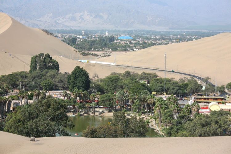 A green lagoon ringed by palms and buildings in a bowl of pale sand dunes, with a city beyond