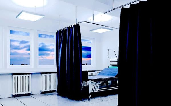 A hospital room with a bed behind a curtain. There are three radiators along the wall underneath a set of windows that show a cloudy sunset outside.
