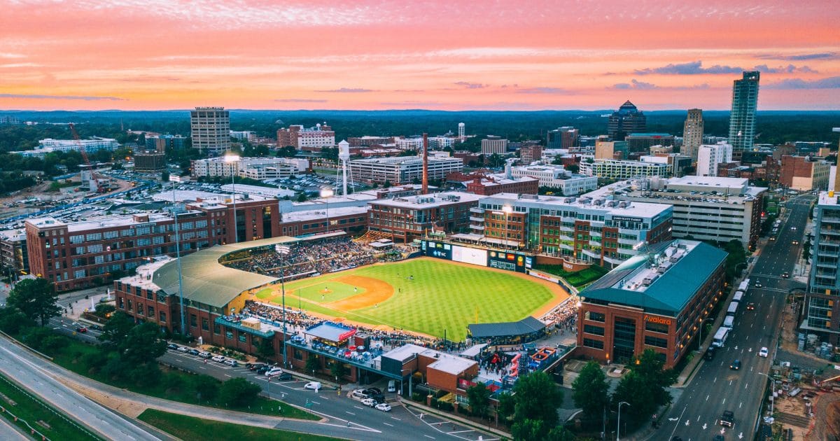 Aerial Image of Durham Bulls Athletic Park and City. Ballpark is centered, game is happening with lights on as the sun sets. City is in the background. 