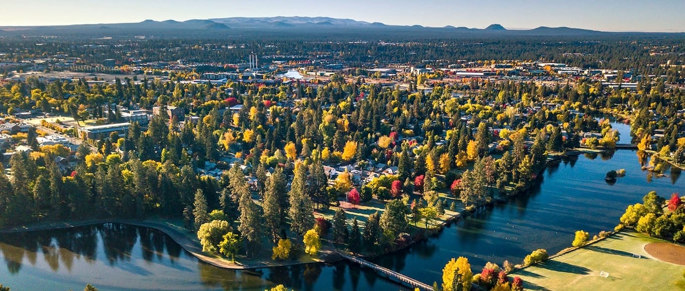 Aerial View of Bend, Oregon. Foreground of image is the Deschutes River. Tall Firs lead to pockets of commercial areas with the City of Bend in the Background. 