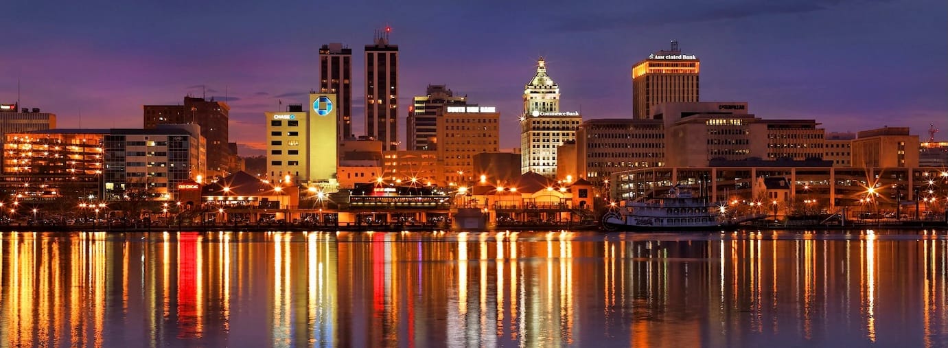 Nighttime Skyline of Peoria, Illinois. The Illinois River is in the foreground and the reflections of the skyline of the city are reflected in the water. The city buildings are lit and appear in the background. 