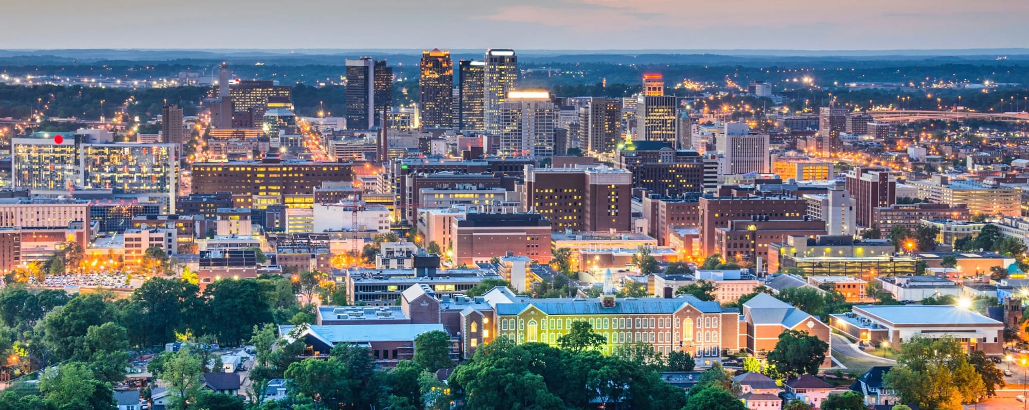 Skyline of Birmingham, Alabama at Dusk. Sun is setting and building lights take over for providing light. Some greenery in the foreground is overshadowed by the downtown buildings in the center and background. 