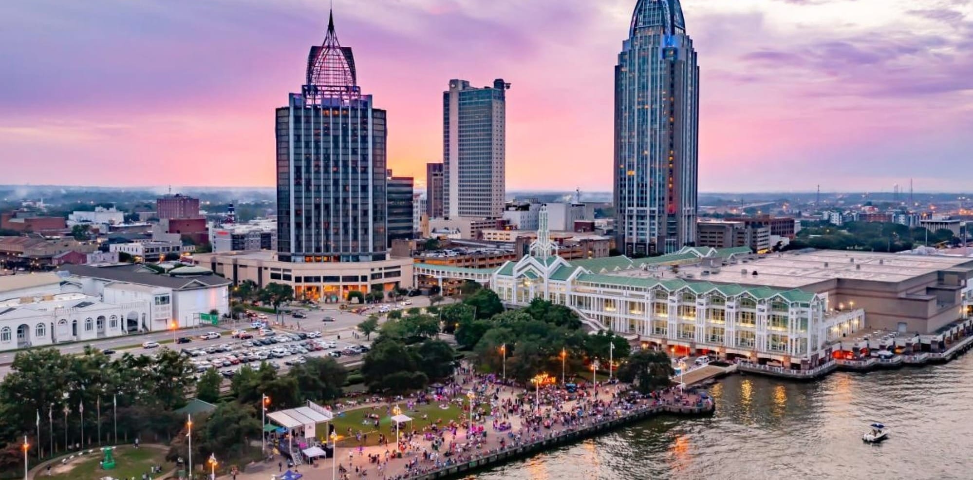 Aerial View of Mobile, Alabama with the Mobile River in the foreground. A summer celebration is happening on the shore of the river and the tall buildings of the city are in the center and background. 