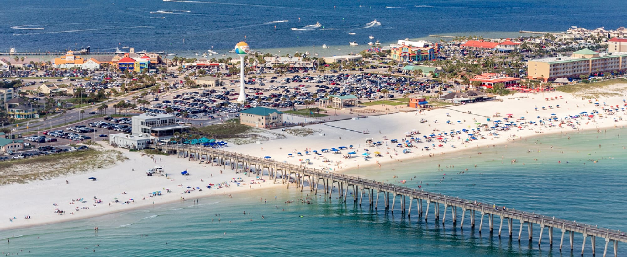 Aerial View of Pensacola Beach on a sunny day. The Beach is full as is the parking lot behind the beach. The gulf pier reaches out from the beach. 