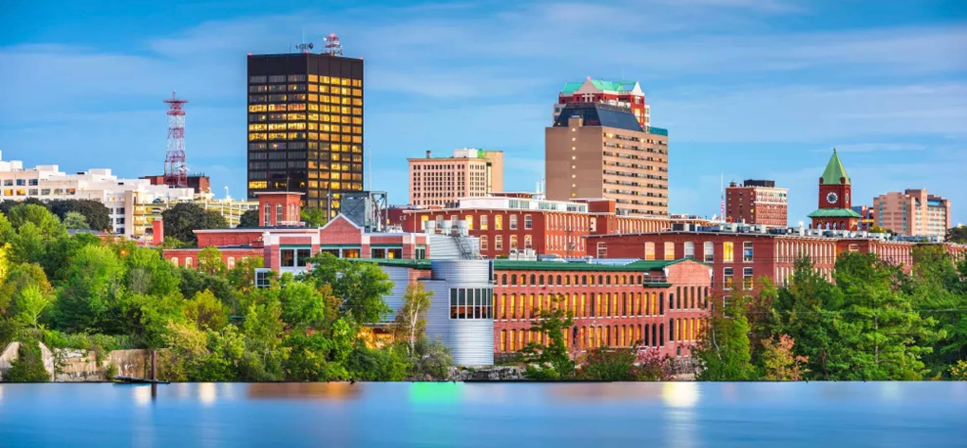 Sunset Image of the Skyline of Manchester, New Hampshire. Beautiful historic brick buildings line the shore of the Merrimack River which is in the foreground. Taller, downtown buildings are in back of the skyline image. 