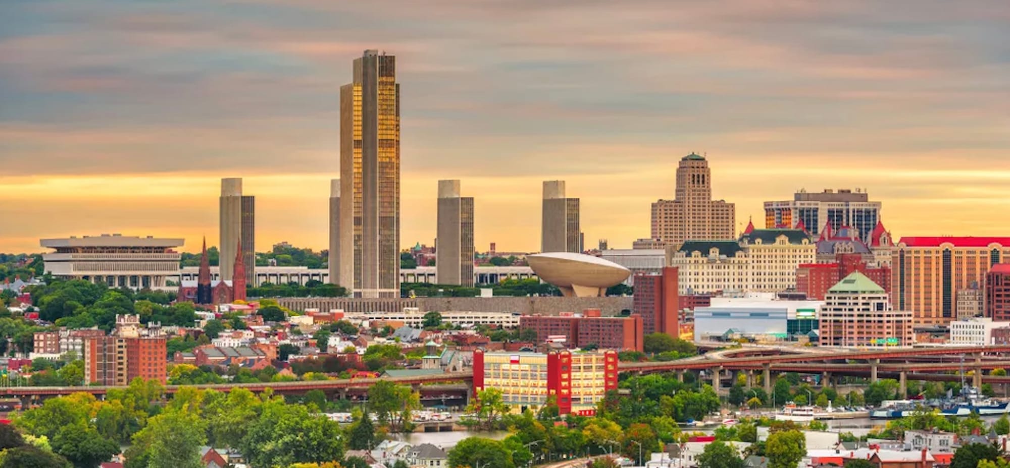 Skyline of Albany New York at Dusk. The Egg, performing arts center building is prominent in the center of the image with taller buildings in the background and light industrial looking buildings in the foreground. 