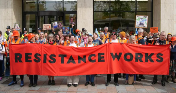 Just Stop Oil protesters hold up an orange banner with black writing on it that says "Resistance Works"