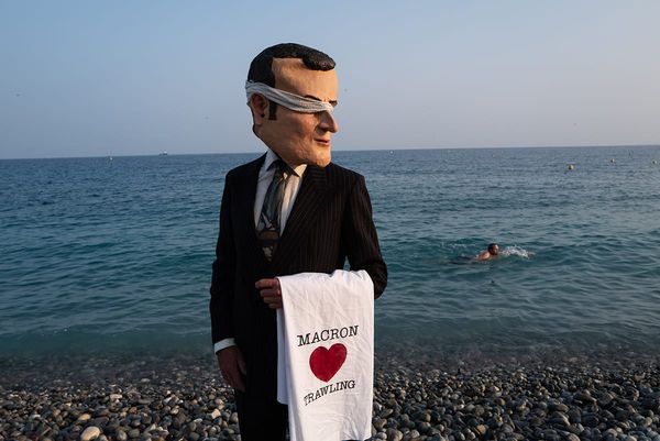 A figure dressed in a suit with an oversize head made of papier mache to represent Emmanuel Macron, holding a t-towel with Macron Heart Trawling written, on the Nice beach