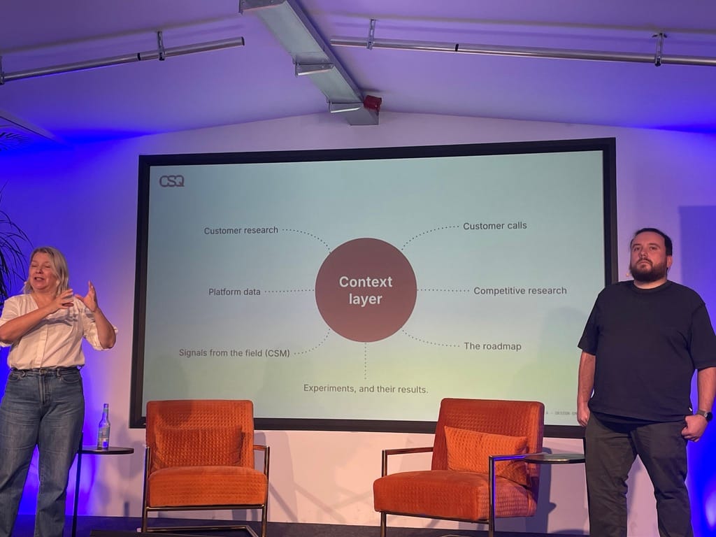 Image of the speakers standing next to their slide, which shows the context layer, including customer research, platform data, signals from the field, experiments and their results, customer calls, competitive research, and the roadmap