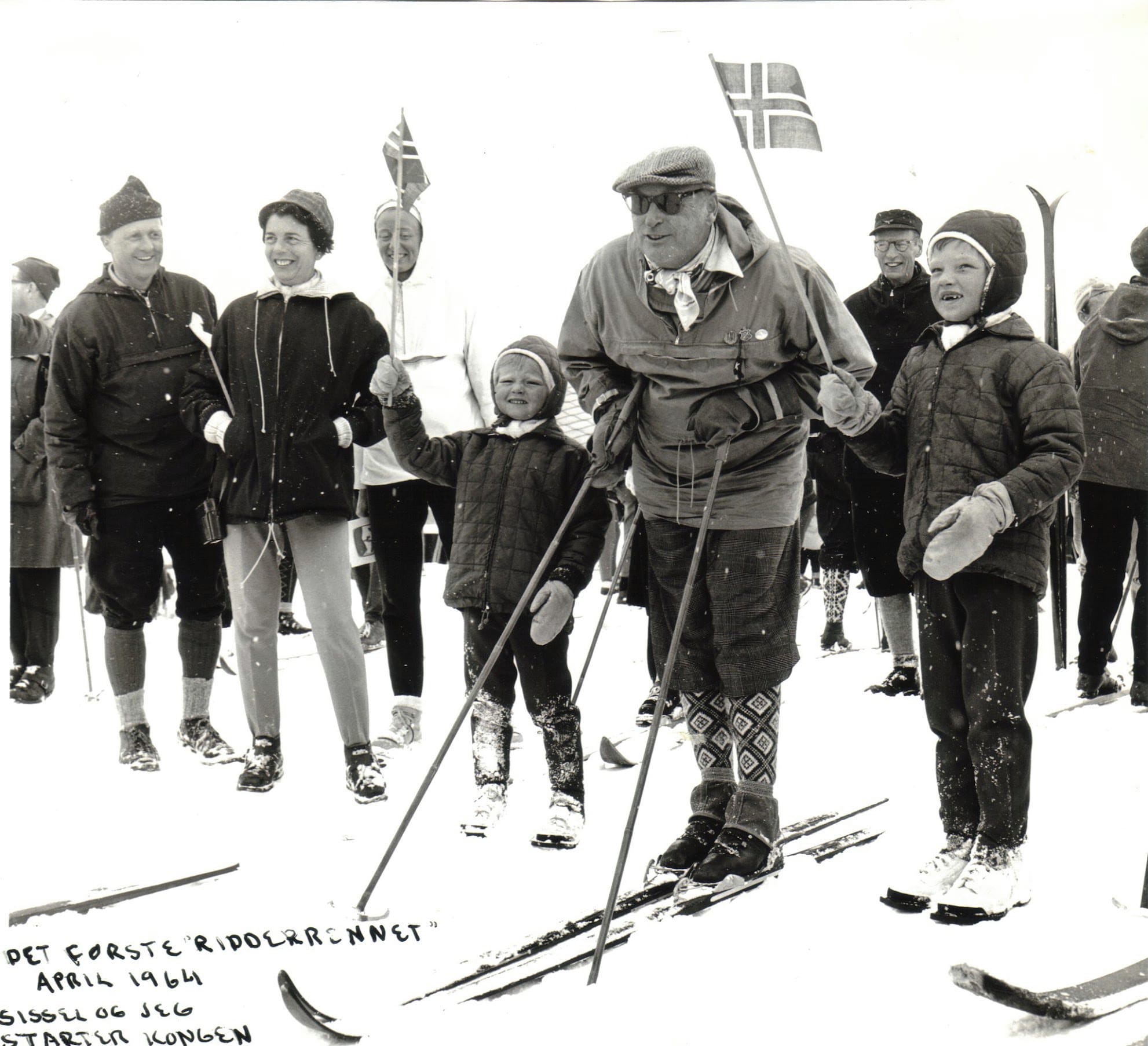King Olav V on skis at the first Ridderrennet, April 1964, flanked by two young children holding small Norwegian flags. A larger Norwegian flag and crowd stand behind him. 