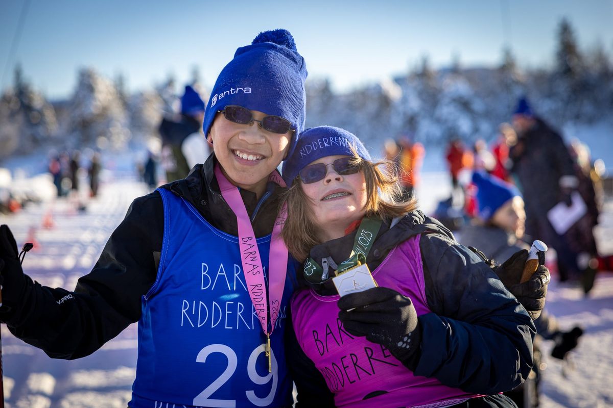 Two smiling children in blue Ridderrennet beanies and sunglasses hug on a sunny winter day, wearing Children's Ridderweek race bibs. One proudly holds up a medal.