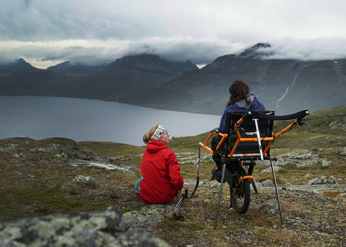 Two people on a rocky plateau above a fjord, with cloud-capped peaks beyond. A woman in a red jacket sits on a stone beside her companion in an orange Joelette off-road wheelchair.