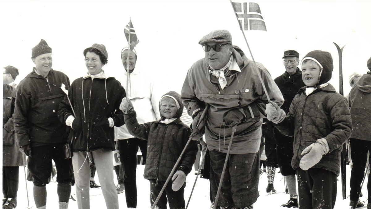 King Olav V on skis at the first Ridderrennet, April 1964, flanked by two young children holding small Norwegian flags. A larger Norwegian flag and crowd stand behind him.