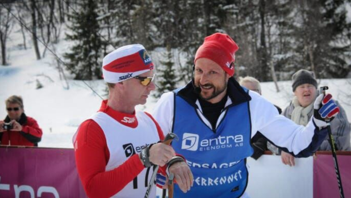 Crown Prince Haakon, in a blue "Ledsager" guide vest and red beanie, stands speaking with a blind elite skier in a red-and-white Swix racing suit. Snowy forest behind them. 