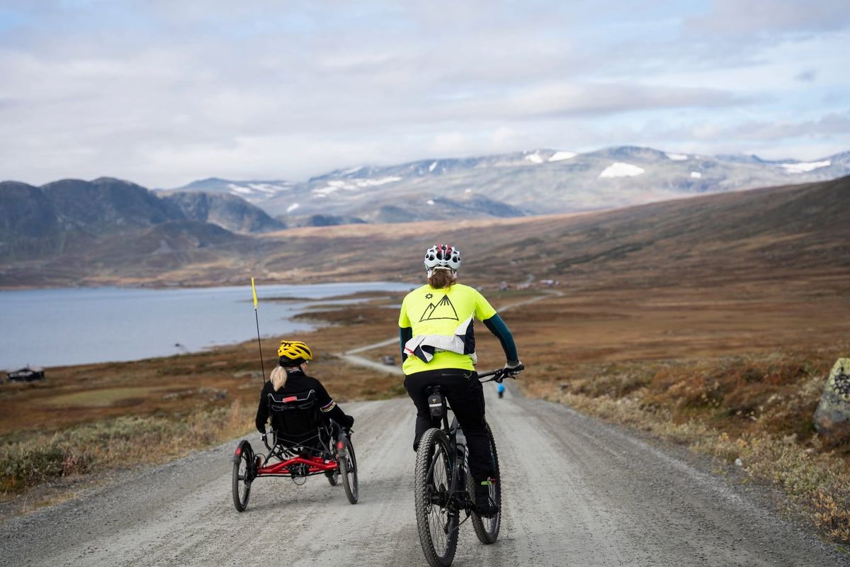Two cyclists, one on a standard mountain bike and one on a red recumbent trike, on a gravel road in an open mountain landscape with a lake in the background.