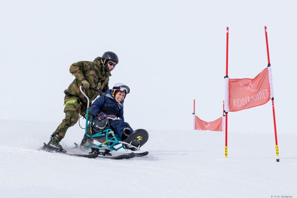 A smiling woman in a sit-ski is guided down a snowy slope by a soldier in Norwegian camouflage uniform. They pass a red Ridderrennet race gate.