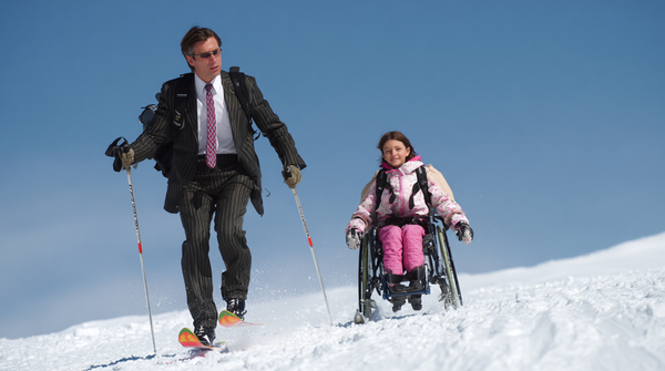 A man in a pinstripe suit and sunglasses skis down a snowy slope beside a young girl in a pink jacket seated in a wheelchair fitted with skis, under a bright blue sky.