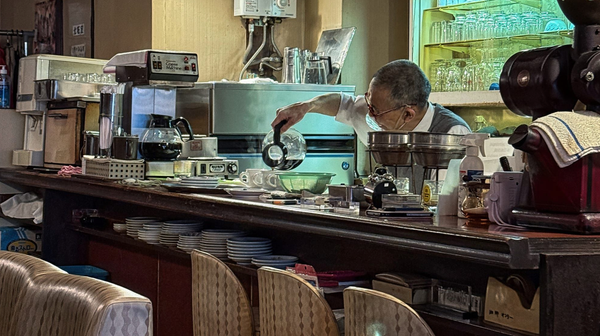 An elderly Japanese barista pours coffee behind the counter of a traditional kissaten, surrounded by vintage cups, glassware and well-worn café equipment.