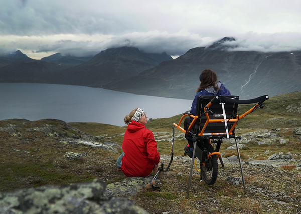 Two people on a rocky plateau above a fjord, with cloud-capped peaks beyond. A woman in a red jacket sits on a stone beside her companion in an orange Joelette off-road wheelchair.