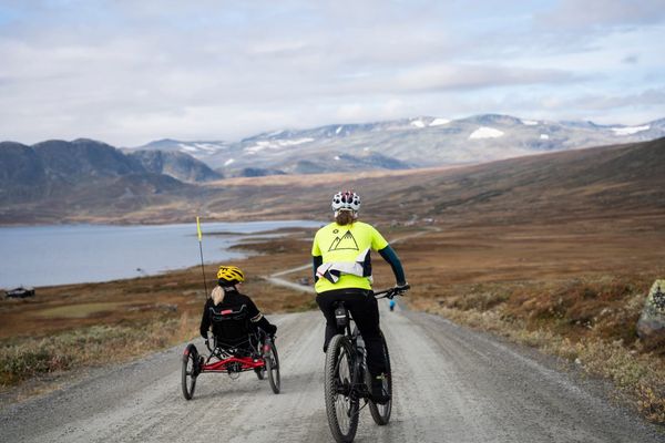Two cyclists, one on a standard mountain bike and one on a red recumbent trike, on a gravel road in an open mountain landscape with a lake in the background.