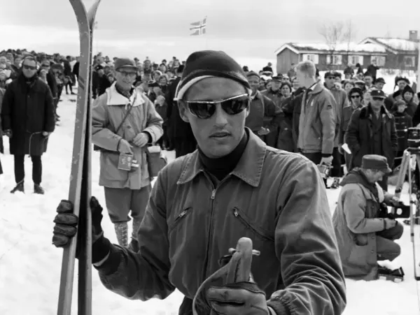 Crown Prince Harald in a dark anorak, ski cap and sunglasses, holding his skis. Behind him a large crowd in the snow, a Norwegian flag and a press photographer kneeling with a camera.