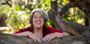 Denise Hancock, a woman, is smiling and leaning over a tree branch