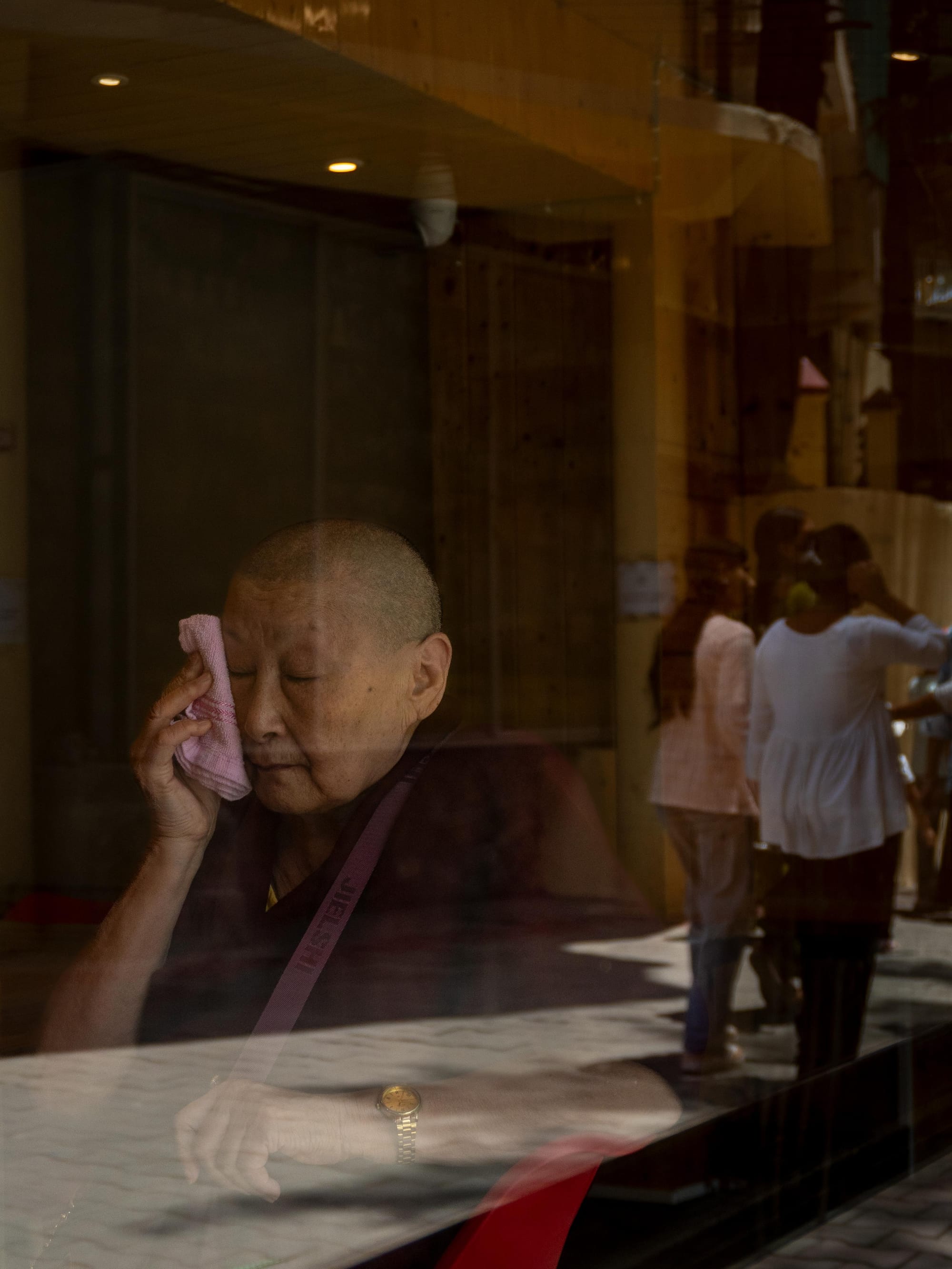 An elderly woman seen through a reflective glass window, with the movement of daily life mirrored on the surface