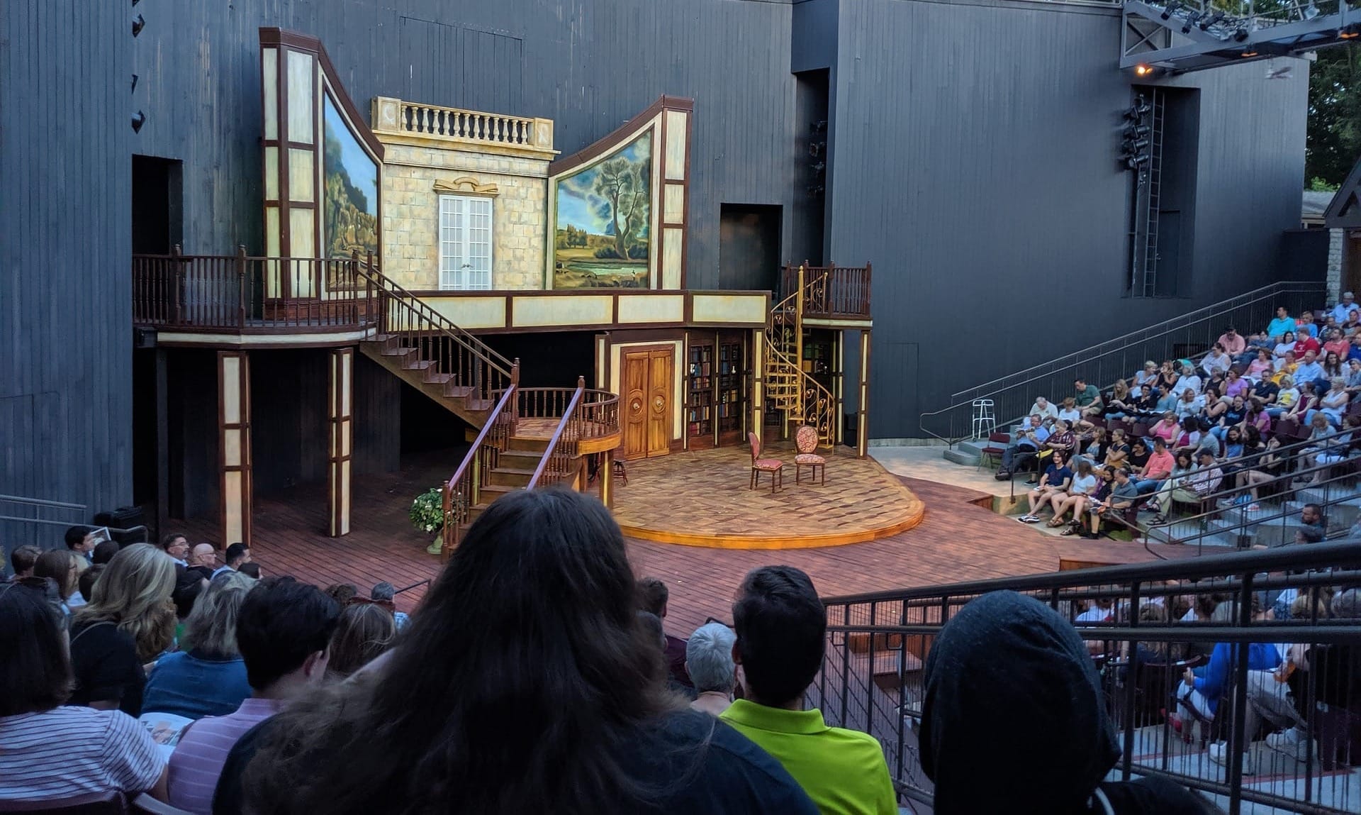 Audience looking at theatre stage on which scenery has been constructed to give the impression that the play is taking place within a grand house.